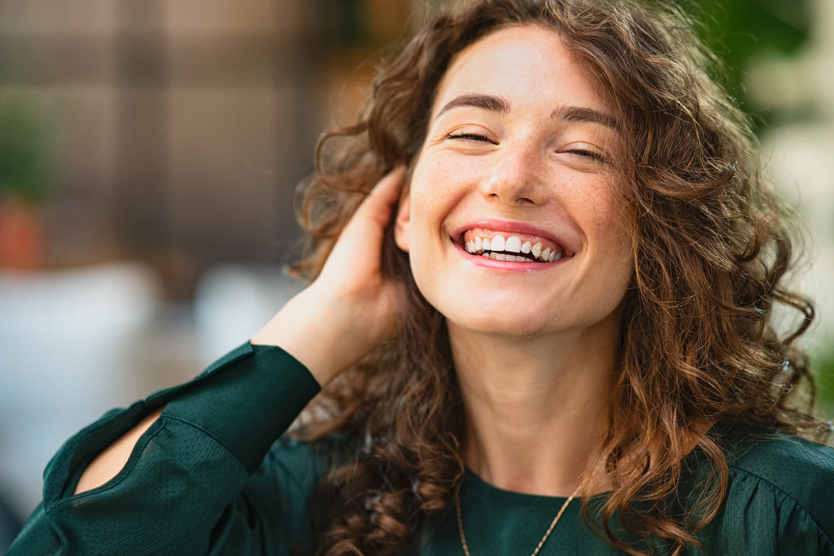 Woman with curly brown hair wearing a dark green shirt grinning at the camera with her hand in her hair