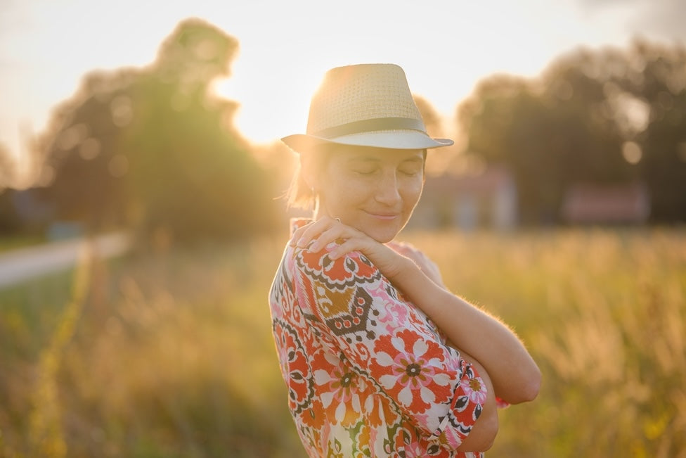 Woman in colourful dress wearing a straw hat with her arms crossed as she smiles in the sunshine