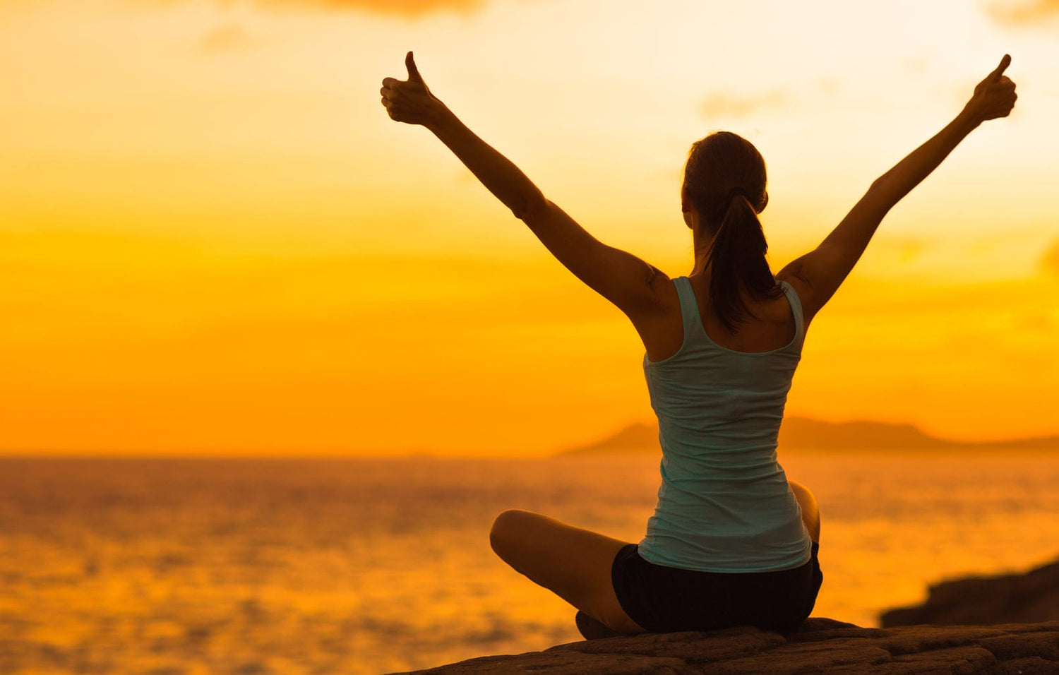 Woman sitting on a cliff side with her thumbs up, arms in the air enjoying the sunset