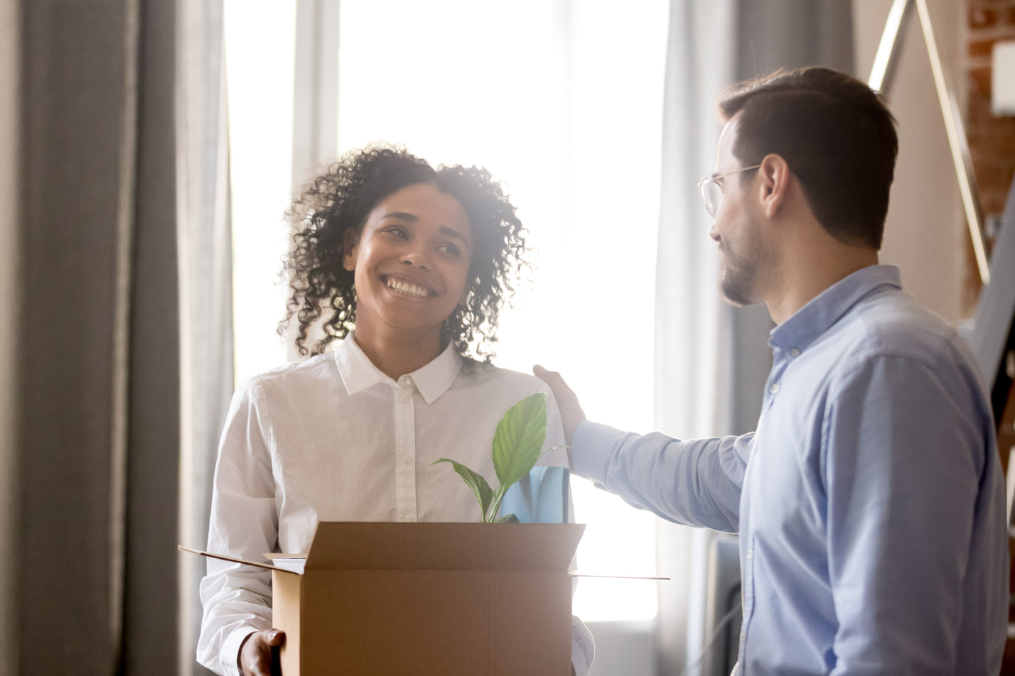 Woman carrying a box of things grinning at man whilst his hand is on her shoulder